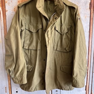 May include: Olive green military-style jacket with multiple pockets, a zipper closure, and button details. The jacket has a high collar and epaulettes on the shoulders. A small emblem is visible on the upper arm. The jacket is hanging on a wooden hanger.