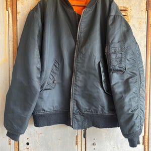 Puede incluir: Una chaqueta bomber azul oscuro con cuello y puños de canalé negros. La chaqueta tiene cierre de cremallera y un bolsillo en la manga izquierda. El forro es naranja.