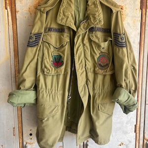 Op de afbeelding: Een olijfgroene militaire jas van de US Air Force met meerdere zakken, patches en de naam "Reynolds" geborduurd op de borst. De jas heeft een ritssluiting en opgerolde mouwen, met een lichtere groene voering. De jas hangt aan een houten hanger.