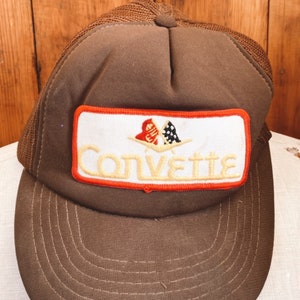 May include: Brown mesh trucker hat with a white patch that says "Corvette" in yellow lettering. The patch also features a red and white checkered flag and a red and white logo.