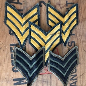 May include: A collection of vintage military chevrons. The chevrons are in two styles: one with yellow and black stripes, and another with green and black stripes. The patches are arranged on a wooden surface.