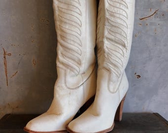 May include: A pair of white leather cowboy boots with a stitched design. The boots have a high heel and are sitting on a metal toolbox.