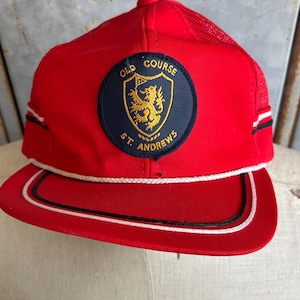 May include: A red baseball cap with a mesh back and a curved brim. The cap features a circular patch with the words "OLD COURSE ST. ANDREWS" and a gold emblem. White and black stripes accent the sides.