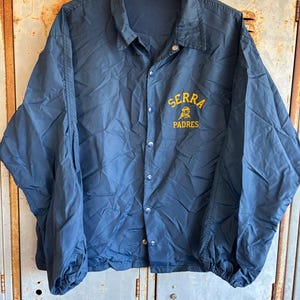 May include: Navy blue nylon jacket with snap closures and a collar. The jacket features the text "SERRA PADRES" in yellow script on the chest. The jacket has elastic cuffs and a drawstring hem.
