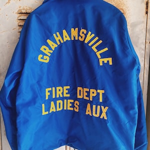 Puede incluir: Chaqueta azul real con letras amarillas. El texto en la parte posterior dice "GRAHAMSVILLE" en arco, con "FIRE DEPT LADIES AUX" debajo. La chaqueta está colgada en una superficie metálica desgastada.