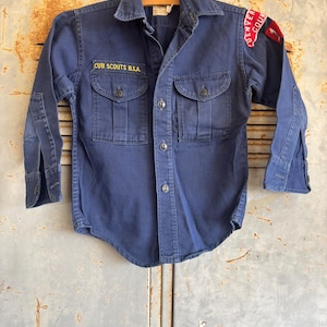 May include: A vintage blue denim Cub Scout shirt with a yellow embroidered "CUB SCOUTS B.S.A." patch on the left chest and a red and yellow embroidered "DENVER COUNCIL" patch on the right shoulder.