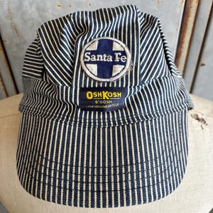 May include: A blue and white striped railroad cap featuring the Santa Fe logo and OshKosh B'Gosh branding. The cap has a rounded top and a short brim, made of a striped fabric.