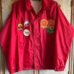 Op de afbeelding: Een felrode nylon jas met een knoopsluiting en een trekkoord aan de onderkant. De jas heeft verschillende patches, waaronder een cactus, een berglandschap en een badge met het nummer 853. De jas hangt aan een houten kleerhanger.