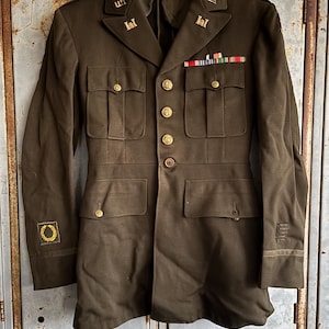 May include: Olive green military jacket with gold buttons, shoulder insignia, and ribbons. The jacket has four front pockets and a tailored fit. The collar features "U.S." lettering. The jacket is hanging on a wooden hanger.