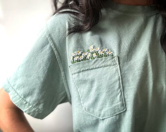 May include: A light green t-shirt with a pocket featuring embroidered white daisies.