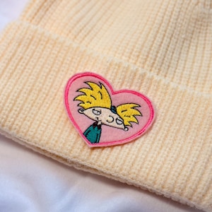 May include: A cream-colored knit beanie featuring a pink heart-shaped patch with an embroidered cartoon character. The character has yellow hair, a green shirt, and a smiling face. The beanie is made of a soft, ribbed knit material.