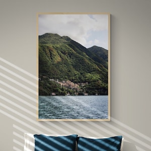 May include: A white bed with a white duvet cover and two blue and orange throw pillows. The bed is in a room with a framed photograph of a lake and mountains hanging on the wall.