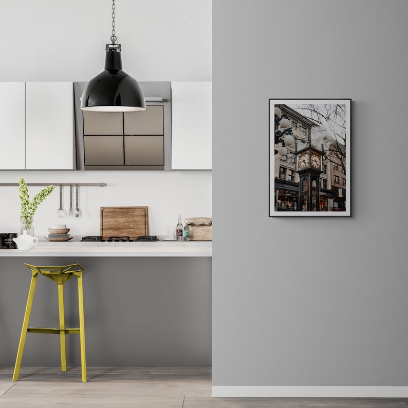 K&ouml;nnte beinhalten: Eine schwarze Pendelleuchte h&auml;ngt &uuml;ber einer K&uuml;chentheke mit einem gelben Hocker. An der Wand h&auml;ngt ein gerahmtes Foto eines Uhrturms in einer Stadt.