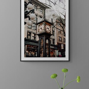 K&ouml;nnte beinhalten: Ein schwarz gerahmtes Foto eines Uhrturms in einer Stadtstra&szlig;e. Der Uhrturm ist aus schwarzem Metall und hat ein goldenes Zifferblatt. Der Uhrturm ist von Geb&auml;uden und B&auml;umen umgeben. Aus einem Schornstein eines der Geb&auml;ude steigt Dampf auf.