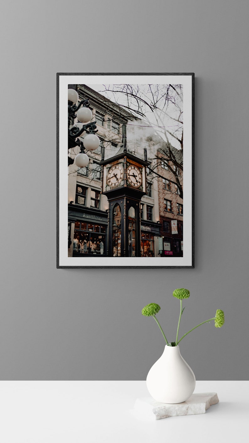 K&ouml;nnte beinhalten: Ein schwarz gerahmtes Foto eines Uhrturms in einer Stadtstra&szlig;e. Der Uhrturm ist aus schwarzem Metall und hat ein goldenes Zifferblatt. Der Uhrturm ist von Geb&auml;uden und B&auml;umen umgeben. Aus einem Schornstein eines der Geb&auml;ude steigt Dampf auf.