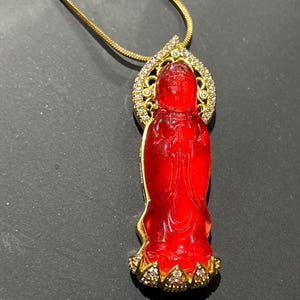 May include: A pendant necklace featuring a translucent red carved figure of Guanyin, set in a gold-toned frame adorned with small clear stones. The figure stands on a lotus base, and the pendant hangs from a thin gold-toned snake chain.