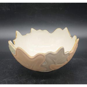 May include: A decorative ceramic bowl with a leaf-like design. The bowl features a cream-colored interior and is adorned with a gold trim along the rim. The exterior is decorated with a pattern of pink, gray, and green leaves.