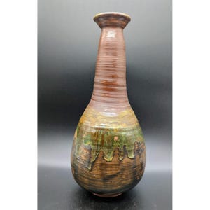 May include: A tall, handcrafted ceramic vase with a brown, green, and tan color scheme. The vase has a textured, ribbed neck and a bulbous body with a dripped glaze effect. The vase is approximately 12 inches tall.