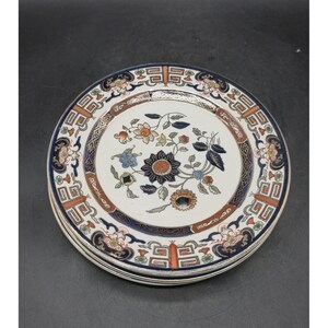 May include: A stack of round ceramic plates featuring a central floral motif in dark blue, orange, and green. The wide rim is decorated with a repeating geometric pattern in dark blue and orange, set against a cream-colored background.