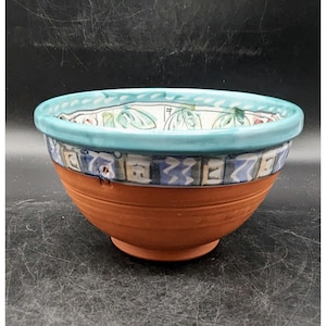 May include: A terracotta bowl with a teal rim and a white, blue, and green patterned design. The bowl has a slightly flared rim and a rounded base.