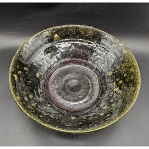 Peut inclure: Un bol en céramique rond avec un vernis brillant sombre présentant des tons tachetés noirs et vert olive. La surface intérieure a un aspect texturé et irrégulier avec un centre profond et sombre et un large bord incliné vers l'extérieur.