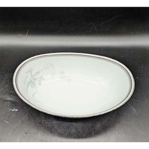 May include: Oval-shaped serving bowl with a white interior and a pale green floral design. The rim features a thin maroon and silver border. The bowl is set against a dark background.