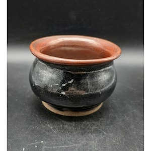 May include: A small, round ceramic bowl with a glossy black exterior finish and a contrasting smooth, reddish-brown rim. The vessel features a wide, open mouth and a slightly tapered base, showcasing a distinct two-tone color scheme throughout.