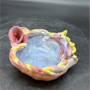 May include: A small ceramic bowl with a pink flower and a pink and yellow bird on the rim. The bowl is glazed in shades of blue, pink, and yellow.