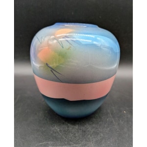 May include: A rounded glass vase featuring a gradient of light blue at the top, a solid horizontal band of soft pink across the center, and a deep blue base. The upper section includes subtle, abstract brushstroke patterns in peach and green.