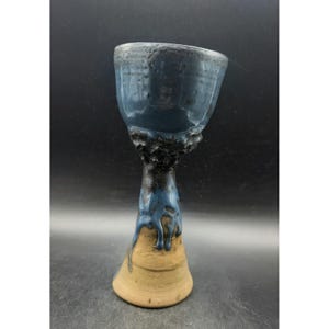 May include: A handmade ceramic goblet with a blue and black glazed exterior. The goblet has a wide, textured rim and a tall, slender stem. The base is a light brown color. The goblet is approximately 6 inches tall.
