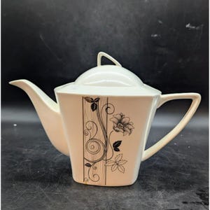 May include: White ceramic teapot with a black and gold floral design. The teapot has a square body and a lid.