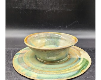 May include: A handmade ceramic bowl and saucer set. The bowl is a light green with a brown rim, and the saucer has concentric rings of green, brown, and yellow. The set is glazed and has a rustic, artisanal appearance.