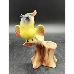 May include: A vintage ceramic bird figurine in yellow, black, and pink, perched on a tree stump planter. The bird has outstretched wings and a detailed design, perfect for home decor.
