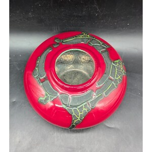 May include: A red ceramic vase with a circular shape and a central opening. The vase features a black and green abstract design around the rim. The interior of the vase appears to be a metallic silver color.