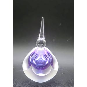 May include: A clear glass perfume bottle with a long, pointed stopper. The bottle has a bulbous shape with a purple interior and a frosted, layered exterior. The design is elegant and decorative.