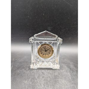 May include: A clear crystal quartz clock with a gold-colored clock face. The clock has a rectangular shape with a triangular top and a diamond-cut pattern. The clock face has black hands and hour markers.
