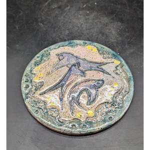 May include: A round ceramic coaster featuring a blue and white design of two stylised fish swimming in a circle. The background is a light pink with yellow dots.