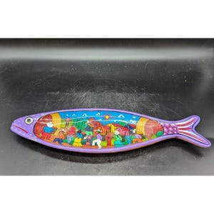 May include: A purple ceramic fish-shaped serving dish with a colorful painted scene of a village with houses and people.