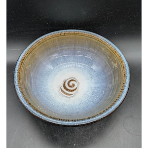 May include: A round ceramic bowl featuring a reactive glaze with a gradient of colors. The center displays a white base with a brown spiral pattern, transitioning into shades of light blue and tan, with a dark brown rim along the outer edge.
