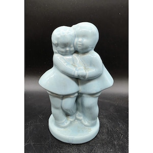 May include: A light blue ceramic figurine depicting two embracing figures. The figures are stylized with rounded features and wear matching outfits. The figurine has a glossy finish and sits on a circular base. The overall design is simple and charming.