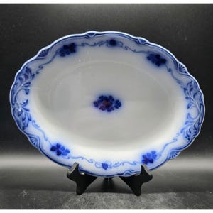 May include: Oval white porcelain serving dish with a blue floral design. The rim has a scalloped edge with blue floral and scrollwork. The center of the dish features a cluster of blue flowers. The dish is displayed on a black stand.