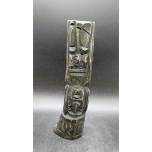 May include: A tall, vertical wooden sculpture featuring hand-carved geometric and abstract facial features. The wood is stained a dark black with lighter, distressed highlights on the raised edges, showcasing a textured, rustic, and weathered finish.