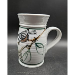 May include: A white ceramic mug with a cylindrical body and a curved handle. The exterior features a hand-painted design of a small bird perched on a brown branch with green leaves. A thin green horizontal line encircles the top and bottom.