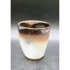 May include: A handmade ceramic tumbler with a gradient glaze. The top is dark brown, transitioning to a tan and then a white base. The interior is dark brown. The cup has a smooth, glossy finish.