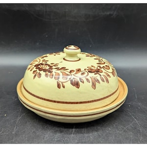 May include: A ceramic butter dish with a lid. The dish is light yellow with brown floral patterns and a matching knob on the lid. The base has two brown stripes. The dish is designed for serving butter.
