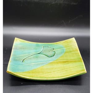 May include: A square ceramic plate with a green and blue glaze. The plate has a raised edge and a subtle curve. A stylized ginkgo leaf is painted in the center of the plate.