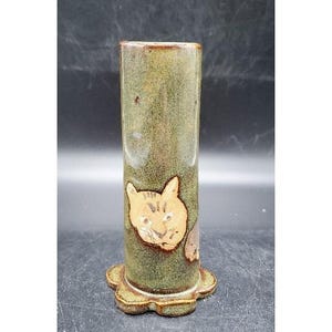 May include: A tall, cylindrical ceramic vase with a cat face design. The vase has a green and brown mottled glaze and a scalloped base. The cat face is tan and brown, adding a whimsical touch to the piece.
