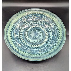 May include: A large, round ceramic bowl with a teal glaze and intricate white designs. The bowl features a swirling pattern of concentric circles and stylized waves.