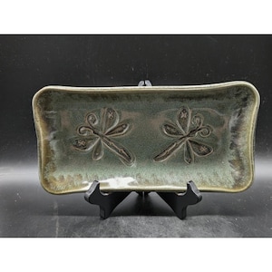 May include: Rectangular ceramic tray with a green and brown glaze. Two dragonfly designs are embossed on the tray's surface. The tray is displayed on a black stand, showcasing its decorative and functional design.