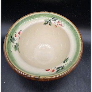 May include: A ceramic bowl with a cream-colored interior and a green rim. The rim is decorated with a brown border and floral accents. The bowl's design includes green leaves and red berries.